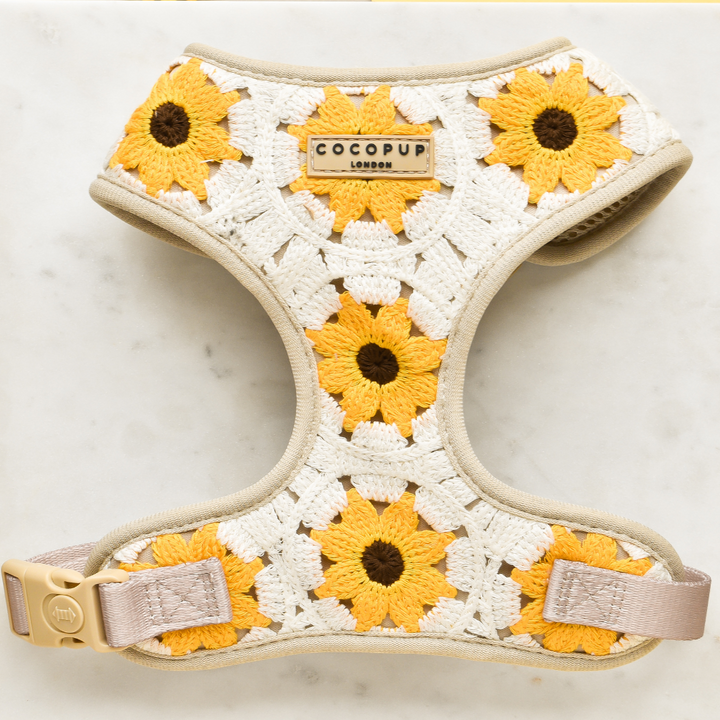 The Sunflower Patch Adjustable Neck Harness by Cocopup London is a beige, stylish dog harness featuring a detailed crochet design adorned with yellow and white sunflowers. It includes a plastic buckle and an adjustable strap. The brand's label is prominently displayed at the center of the harness. The product is presented on a white marble surface.