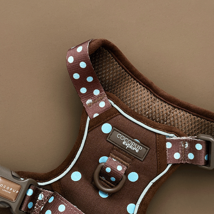 A close up of the Cocopup logo and d-ring on a brown dog harness with light blue polka dots, laid flat on a solid brown background. 