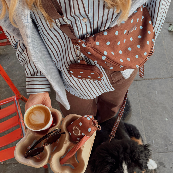 A person wearing a brown and blue polka dot crossbody bag and matching pouch, holding a coffee cup outdoors next to a black dog.