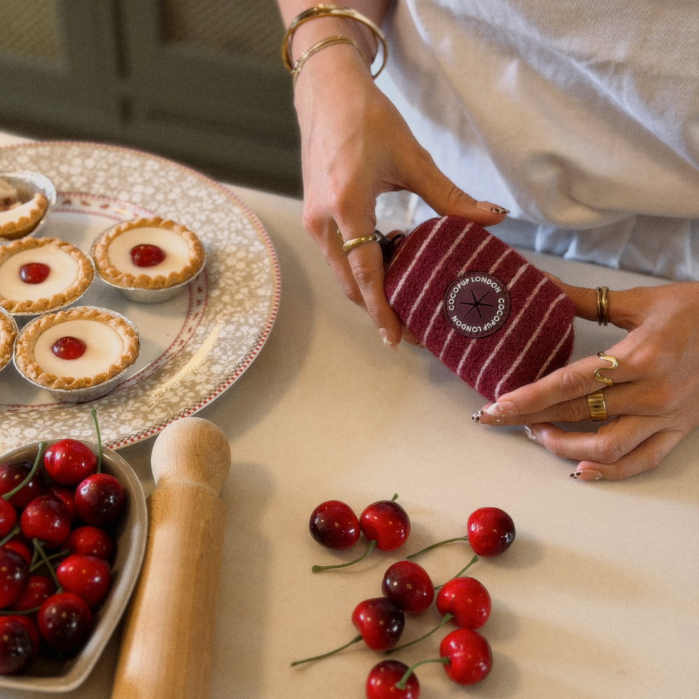 A person holds a small striped fabric poop bag holder with a circular embroidered logo on a beige tabletop. The scene includes cherry pies on a plate, cherries in a bowl, and a wooden rolling pin, indicating a kitchen or bakery setting.