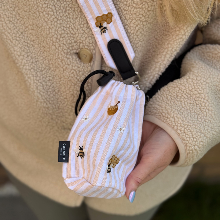 Cocopup London drawstring treat pouch in Honey Meadow, featuring a bees and honeycomb print, worn with a beige coat.