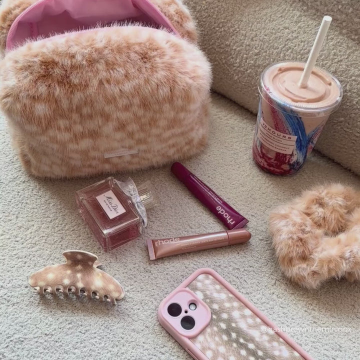 A styled flat-lay of a fluffy faux-fur cosmetic pouch in a warm beige and brown pattern, shown alongside a pink phone case, hair accessories, a perfume bottle, and an iced coffee on a carpeted floor.