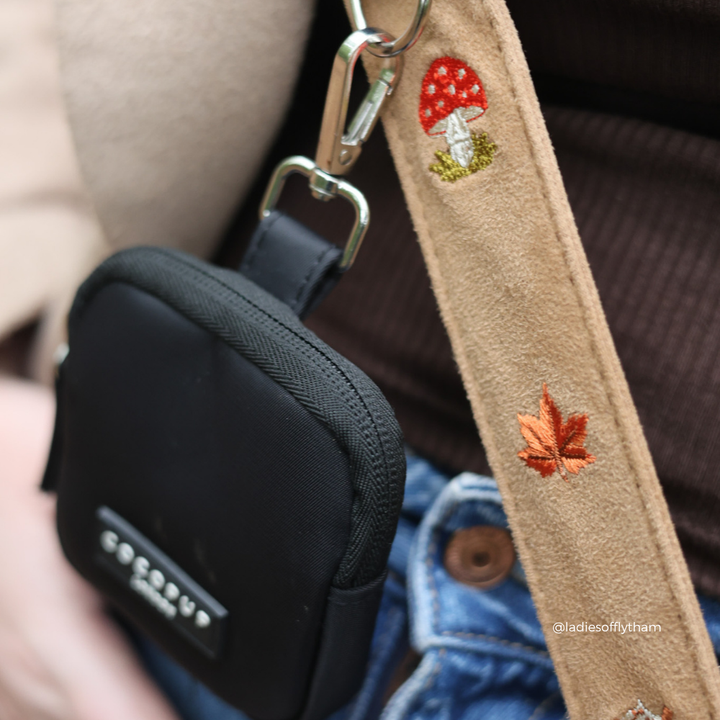 Close-up of a black fabric crossbody treat pouch with a tan suede strap featuring embroidered red and orange mushrooms and a leaf