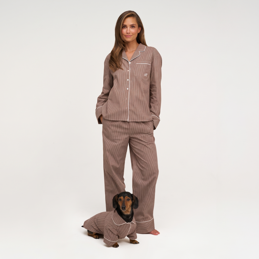 A woman wearing a brown pinstriped pajama set stands barefoot against a white studio background, with a small dachshund dog sitting in front of her wearing a matching pajama top. The pajamas have a warm taupe-brown color with thin white pinstripes and white piping.