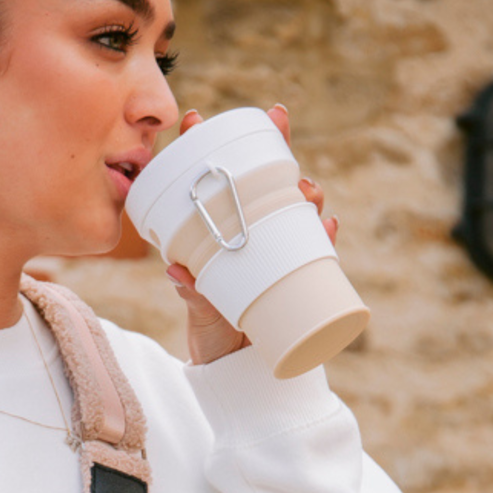 Model drinking from the Plain Nude with White Lid Collapsible Silicone Travel Coffee Cup with Cocopup branding and carabiner clip.